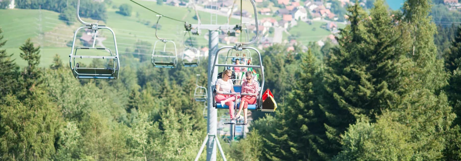 zwei Frauen auf einem Sessellift inmitten von grünen Bäumen.